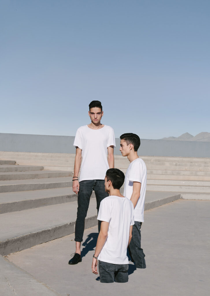 Victor Zepeda's image captures three male figures in white t-shirts against a backdrop of clear blue skies and concrete steps. The tallest man stands prominently in the center while the other two are positioned to his sides creating a sense of depth and perspective.